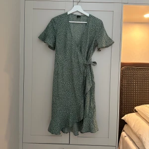 Grön prickig omlottklänning - Fin sommarklänning i omlott. Grön med små vita prickar. Volang nedtill och på ärmarna. Sval och skön. I använt men gott skick. 
