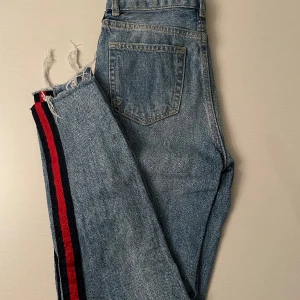 Jeans med röd rand på sidan - Blå jeans (lite ljusare i verkligheten) med röd rand på sidan av benen och lite slitningar. Från bikbok, i bra skick! 