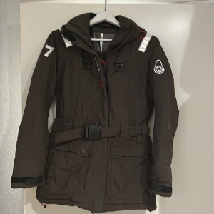 Sail Raising jacka med skärp  - Sail Raising jacka i lite längre modell. Avtagbart midjeskärp. Jättefint skick i trendig brun färg i stl S. Avtagbar luva. Gore Tex. Hör av er för fler bilder. 