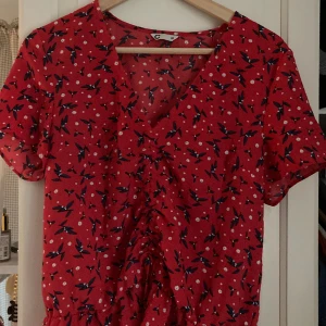 Blus - Kortarmad blus med blå, svart, vit o röda mönster/blommor. Denna är som en magtröja och kan reglera om man vill att magen syns mer. Kan postas eller hämtas i Lund.