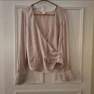Beige omlottblus - En elegant beige omlottblus med långa ärmar och knappar vid ärmsluten. Blusen har en draperad effekt vid midjan och är tillverkad i ett mjukt och lätt material.