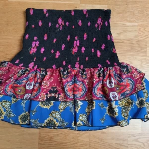 Mönstrad kjol från Neon & Nylon - Snygg och färgglad kjol från Neon & Nylon med flera olika mönster och färger. Kjolen har en svart överdel med rosa prickar, följt av ett rött parti med paisleymönster och ett blått parti med blommönster. Kjolen har en volangdetalj och är av kort längd ✨