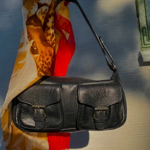 Svart axelväska - En svart axelväska i skinn med två framfickor som har spännen. Väskan har en justerbar axelrem och en dragkedja för att stänga huvudfacket.