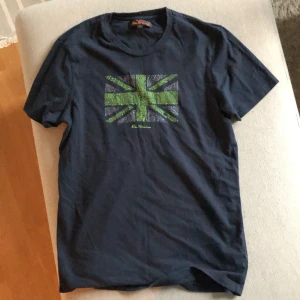 Mörkblå t-shirt från Ben Sherman - En mörkblå t-shirt från Ben Sherman med ett grönt och grått mönster av Union Jack-flaggan på framsidan. T-shirten har korta ärmar och en normal passform. Tröjan har aldrig använts och är i nyskick. Säljs billigt då jag vill bli av med det!
