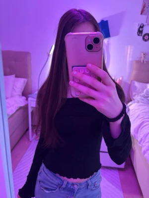 Svart croppad långärmad topp - Säljer en svart croppad topp med långa ärmar och lätt vågig kant nertill. Toppen har en slim passform och är perfekt att styla med höga jeans. Materialet känns mjukt och stretchigt, troligen bomull eller polyester.