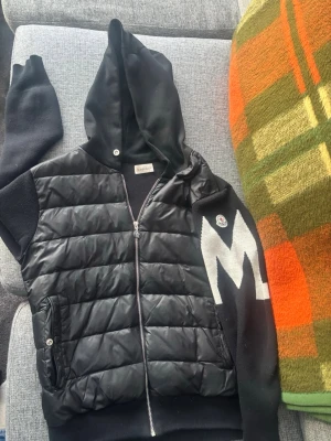 Svart Moncler hoodie med dragkedja man måste dra upp drag kedjan försiktigt annars så uppnas den lite men då är de bara och gö om de går säkert och fixa. Nypris:12000 kr storlek:M  - Snygg svart hoodie från Moncler med quiltad front och stickade ärmar. Stor vit M-detalj på ena ärmen och klassisk Moncler-logga. Dragkedja framtill, två fickor och stor huva. Materialmix av dun/quiltad polyester och mjuk bomull. Perfekt för en trendig streetwear-look.