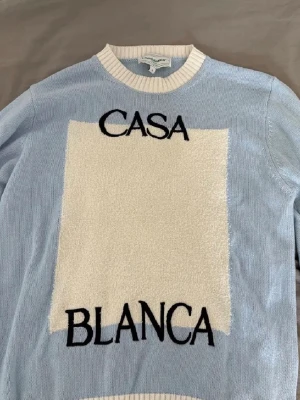 Ljusblå stickad tröja Casablanca - Snygg ljusblå stickad tröja från Casablanca med vit, fluffig panel framtill och svart broderad text 'CASA BLANCA'. Tröjan har ribbstickad vit krage och är perfekt för dig som gillar trendiga detaljer och streetwear-vibe.
