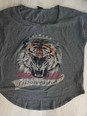 Grå t-shirt med tigertryck Gina Tricot - Cool grå t-shirt från Gina Tricot i storlek XS med ett stort tigertryck framtill och textdetaljer i rött och silver. T-shirten har rund halsringning och korta ärmar. Perfekt för dig som vill sticka ut med ett djurmotiv.