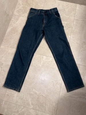 Mörkblå Carhartt jeans straight fit - Säljer ett par mörkblå jeans från Carhartt med raka ben och klassisk femficksdesign. Jeansen har extra fickor på sidorna och bak, samt Carhartt-logga på ena bakfickan. Tillverkade i slitstark denim, perfekta för en avslappnad streetwear-look. 30/34 men passar allt mellan 28/32 till 32/36