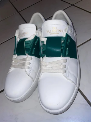 Valentino Garavani vita sneakers - Snygga vita sneakers från Valentino Garavani med gröna detaljer och vita skosnören. Skorna har en klassisk låg siluett och är tillverkade i slätt läder. Perfekta för dig som vill ha en clean och exklusiv look med en twist. Har bara använt drygt 1-5 gånger endast så nästan i ny skick, passar 44,43,42