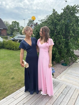 Marinblå balklänning  - Elegant marinblå långklänning med djup v-ringning och korta ärmar. Klänningen har en figurnära passform upptill och faller snyggt längs kroppen. Perfekt för speciella tillfällen och har en klassisk, stilren look. Klänningen har även slits på sidan av benet så det är lätt att gå i! 