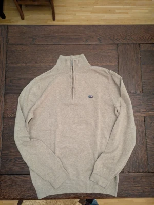 Beige halvzip tröja från Lexington - Snygg beige stickad tröja från Lexington med halvzip och liten logga på bröstet. Tröjan har hög krage, långa ärmar och ribbade muddar vid ärmslut och nederkant. Perfekt för lager på lager och enkel att matcha med jeans eller chinos.