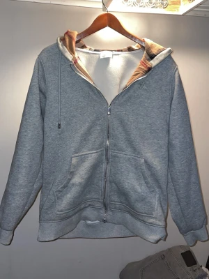 Grå zip hoodie med rutigt foder - Säljer en grå hoodie med dragkedja från Burberry. Hoodien har ett klassiskt rutigt foder i huvan och broderad logga på bröstet. Två stora fickor framtill och justerbar huva med snören. Perfekt för en chill och stilren look.