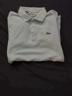 Ljusblå pikétröja från Lacoste - Klassisk ljusblå pikétröja från Lacoste med krage och knappar framtill. Tröjan har det ikoniska gröna krokodilmärket broderat på bröstet. Perfekt för en stilren och avslappnad look.