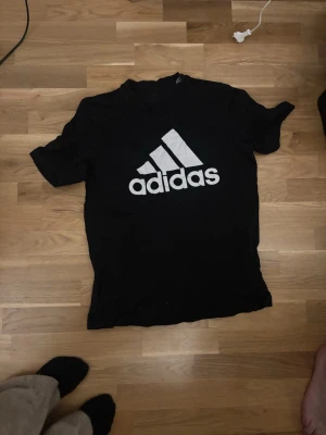 Svart Adidas t-shirt med logga - Svart t-shirt från Adidas med stor vit logga på bröstet. Klassisk passform och rund halsringning, perfekt för en sportig och avslappnad stil. Tillverkad i mjukt material som känns skönt mot huden.