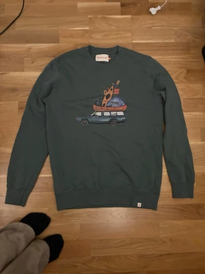 Grön sweatshirt med tryck Revolution - Cool grön sweatshirt från Revolution med ett unikt tryck av en bil med pizza och planet på taket. Tröjan har rund hals, långa ärmar och ribbade muddar. Perfekt för dig som gillar streetwear och snygga prints.