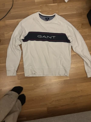Vit sweatshirt från GANT - Vit sweatshirt från GANT med rund halsringning och en bred marinblå rand över bröstet med GANT-loggan broderad i vitt. Tröjan har röda detaljer längs randen och ribbade muddar vid ärmslut och nederkant. Perfekt för en stilren och sportig look.