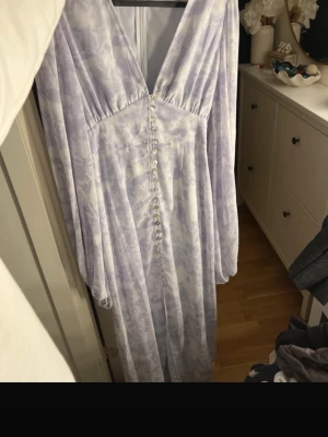 Adoore Positano  - Superfin långklänning i ljuslila och vitt med subtilt mönster. Djup v-ringning, långa ärmar och dekorativa pärlemorknappar framtill. Klänningen har en romantisk och flowy känsla, perfekt för dig som gillar en feminin stil. Finns inte längre att köpa i detta mäster 