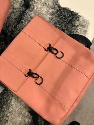 Rosa ryggsäck från Rains - Snygg rosa ryggsäck från Rains i vattenavvisande material med två svarta metallspännen framtill. Ryggsäcken har en stilren, fyrkantig form och justerbara axelband i svart. Perfekt för dig som vill ha en trendig och praktisk väska.