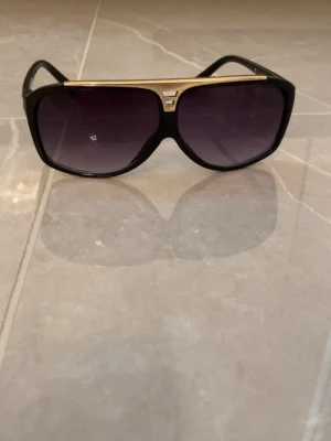 Louis Vuitton svarta solglasögon - Snygga svarta solglasögon från Louis Vuitton med guldiga detaljer och mörka tonade glas. Bågarna har en klassisk aviatorform och loggan är ingraverad på sidan. Perfekta för dig som vill ha en lyxig och trendig accessoar till sommaren.