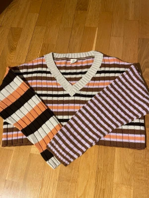 RandigNy stickad tröja Urban Outfitters - Säljer en ribbstickad tröja från Urban Outfitters med breda ränder i brunt, lila, orange, beige och svart. Tröjan har V-ringning och långa ärmar. Perfekt för dig som gillar färgglada och retroinspirerade plagg. Tröjan är ny, aldrig använd. Storlek xs.