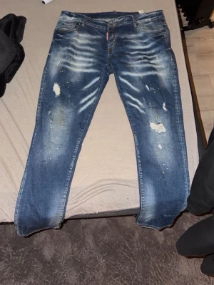 Blå och svarta jeans från dsq2 - Säljer 2 par blå och svarta jeans från Dsq2 med slitna detaljer, färgstänk och tydliga slitningar framtill. Klassisk femficksmodell med gult märke bak i midjan. Jeansen har en rak passform och är tillverkade i denim. Privat för att diskutera om pris sänkning.