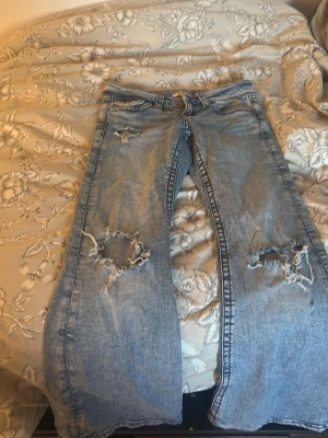 Ljusblå slitna jeans - Säljer ett par ljusblå  jeans med stora slitningar på båda benen och klassiska fem fickor. Jeansen har en avslappnad passform och är tillverkade i denim. Perfekta för en trendig och avslappnad stil.