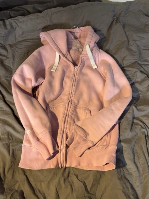 Ljusrosa zip hoodie från Nike - Säljer en ljusrosa hoodie från russemerch  med dragkedja framtill och vita snören i huvan. Hoodien har ribbade muddar vid ärmslut och nederkant samt fickor på sidorna. Perfekt för en avslappnad och sportig stil.
