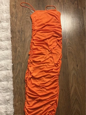 Orange långklänning med smala band - Trendig orange långklänning med smala axelband och rynkade sidor som ger en snygg draperad effekt. Klänningen är figurnära och har en enkel, stilren design som passar perfekt till sommarens alla tillfällen.