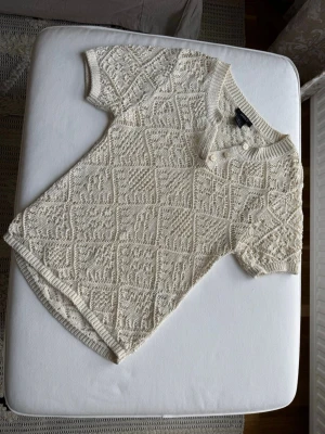 Beige stickad topp med kort ärm - Snygg beige stickad topp med korta ärmar och ribbade kanter. Toppen har ett diskret rutmönster och knappar vid halsringningen för en extra detalj. Perfekt till jeans eller kjol för en avslappnad look.