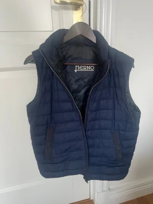 Mörkblå dunväst från Herno - Snygg mörkblå dunväst från Herno med quiltad design och hög krage. Västen har två fickor framtill med dragkedja och en diskret metallogo bak i nacken. Perfekt för lager-på-lager och stilrena outfits.Har även en luva man kan stoppa in och ta ut 