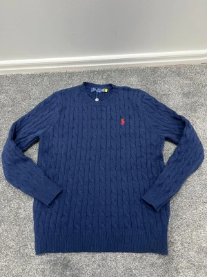 Blå kabelstickad tröja Polo Ralph Lauren - Mörkblå kabelstickad tröja från Polo Ralph Lauren med rund halsringning och röd broderad logga på bröstet. Tröjan har långa ärmar och ribbade muddar vid ärmslut och nederkant. Perfekt för en stilren och klassisk look.