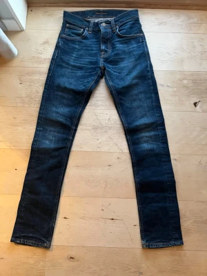Mörkblå jeans från Nudie Jeans - Säljer ett par mörkblå jeans från Nudie Jeans med klassisk femficksmodell och kontrastsömmar. Jeansen har en slim passform och är tillverkade i slitstarkt denim med snygg tvätt och detaljerade bakfickor.