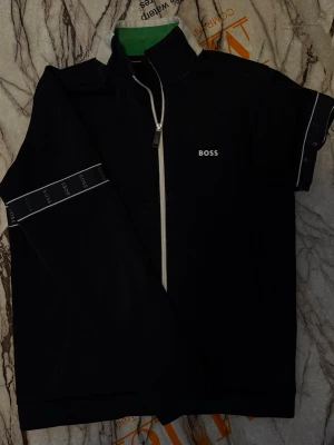 Svart ziptröja från BOSS - Svart långärmad ziptröja från BOSS med vit dragkedja och logga på bröstet. Tröjan har hög krage och dekorativa band med BOSS-tryck längs ärmarna. Perfekt för en stilren och sportig look. Nypris kostade 2499 kr, plus att denna model inte finns längre kvar i deras försäljning. 