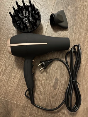 Svart hårfön med tillbehör - Säljer en svart hårfön med matt finish och detaljer i guld. Kommer med två munstycken: en diffuser och en smal fönmunstycke. Utrustad med lång sladd och europeisk kontakt. Perfekt för styling och torkning av hår.