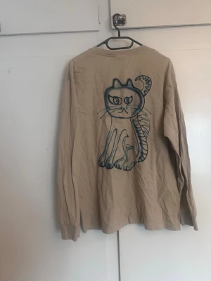 Beige långärmad topp med kattmotiv - Beige långärmad topp från Weekday i bomull med ett stort, tecknat kattmotiv på ryggen. Rund halsringning och avslappnad passform. Perfekt för dig som gillar unika prints och vill sticka ut lite extra.