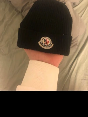 Svart Moncler mössa i ull - Svart ribbstickad mössa från Moncler med klassisk Moncler-logga framtill. Går att fraktas och sänka priset vid en snabb affär. Skriv om ni har några andra frågor 