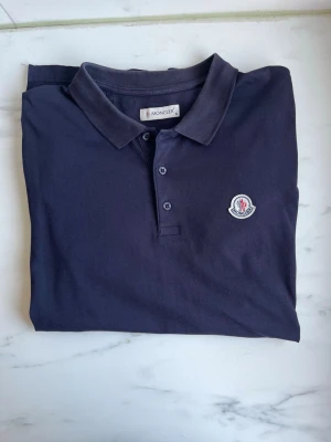 Moncler Pike  - Snygg marinblå pikétröja från Moncler med klassisk krage och knappar framtill. Tröjan har korta ärmar, är storlek XL fits L/M, helt fint skick på tröjan inga defekter ! 