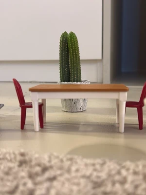 Dockmöbler med bord, stolar och kaktus - Säljer ett set med dockmöbler som inkluderar ett rektangulärt bord med träfärgad topp och vita ben, två röda stolar samt en grön kaktus i vit kruka. Perfekt för dockhus eller miniatyrsamlingar.