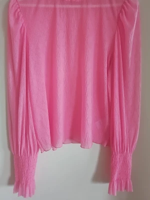 Rosa blus med puffärmar - Trendig rosa blus med lång puffärm och smockade muddar. Tillverkad i ett lätt, strukturerat tyg som ger en luftig känsla. Perfekt för dig som vill sticka ut med färg och volym i garderoben.