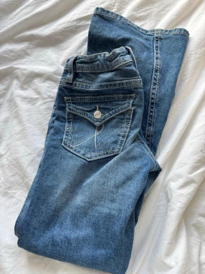 Blå jeans från Gina Tricot - Säljer ett par klassiska blå jeans från Gina Tricot med raka ben och snygga kontrastsömmar. Jeansen har fem fickor, inklusive en bakficka med lock och knappdetalj. Tillverkade i mjukt denimtyg med en tidlös tvätt.
