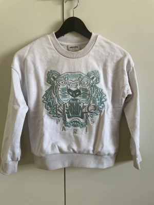 Vit Kenzo Kids sweatshirt med tiger - Säljer en vit sweatshirt från Kenzo Kids med ikoniskt tigerbroderi i grönt och grått framtill. Tröjan har rund hals, ribbade muddar och är tillverkad i mjukt bomullsmaterial. Perfekt för dig som gillar streetstyle och vill sticka ut med en cool logga.