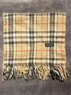 Beige rutig halsduk från Burberrys - Klassisk beige halsduk från Burberrys med ikoniskt rutmönster i brunt, vitt och rött. Tillverkad i 100% lammull och har fransar i ändarna. Perfekt accessoar för att lyfta din outfit med en tidlös brittisk stil.