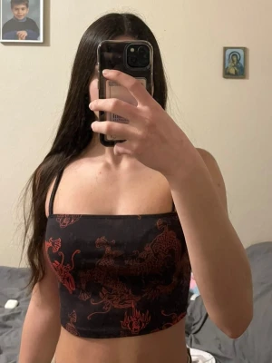 Svart och röd croppad linnetopp - Trendig croppad linnetopp i svart med röda drakmönster. Toppen har smala axelband och sitter tight mot kroppen. Perfekt för dig som vill sticka ut med en cool och edgy stil.