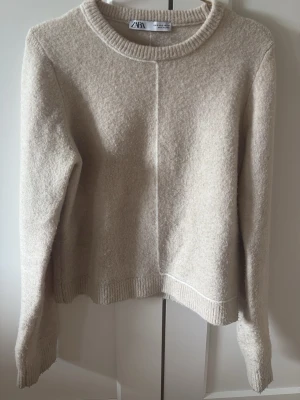 Beige stickad tröja från Zara - Mysig beige stickad tröja från Zara med rund halsringning och synlig söm framtill och baktill. Tröjan har långa ärmar och ribbade muddar vid ärmslut och nederkant. Perfekt för lager på lager och enkel att matcha med olika outfits.