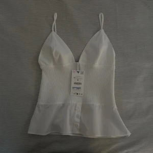 Vit topp från Zara  - En helt ny och snygg vit topp med smala axelband och v-ringning. Toppen har en ribbad mittdel och knappar framtill. Ny pris 329kr