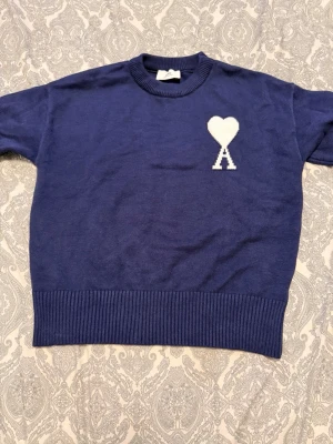 Blå stickad tröja från Ami Paris - Säljer en blå stickad tröja från Ami Paris med deras ikoniska vita hjärta och A-logga på bröstet. Tröjan har rund halsringning, ribbade muddar och är gjord i mjukt material. Perfekt för dig som gillar stilrena och trendiga plagg.
