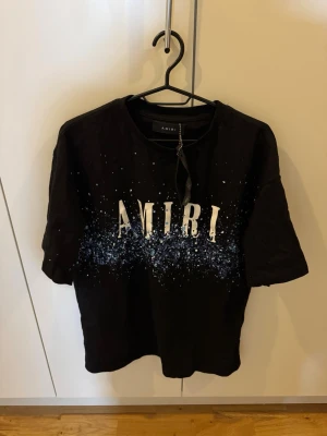 Svart Amiri t-shirt med tryck - Cool svart t-shirt från Amiri med stort vitt logotryck och blått stänkmönster framtill. Klassisk passform med rund hals och korta ärmar. Perfekt för dig som vill sticka ut med en streetstyle-look.