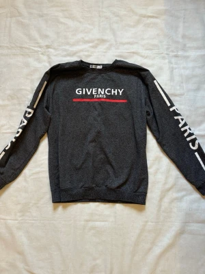 Mörkgrå oäkta Givenchy Paris sweatshirt - Inte äkta - den är köpt utomlands. Köpt från män avdelningen men tog själv på mig den som kvinna. Snygg mörkgrå sweatshirt med vit logga och röd linje på bröstet. Texten 'PARIS' finns även tryckt längs båda ärmarna. Tröjan har rund halsringning och är tillverkad i mjukt material, perfekt för en trendig streetwear-look. Storleken i etiketten är inte i EU. Bedömer att den är i storlek M. Bröstmått är 51cm. Längd är 65cm.