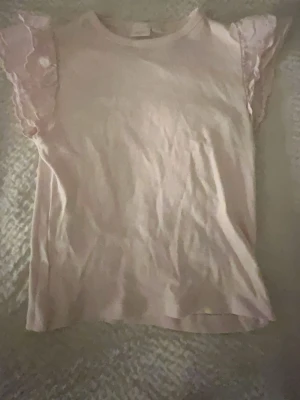 Ljusrosa topp med volangärmar - Supersöt ljusrosa topp med volangdetaljer på ärmarna. Toppen har rund halsringning och är tillverkad i ett mjukt bomullsmaterial. Perfekt för dig som gillar romantiska detaljer och en feminin stil.